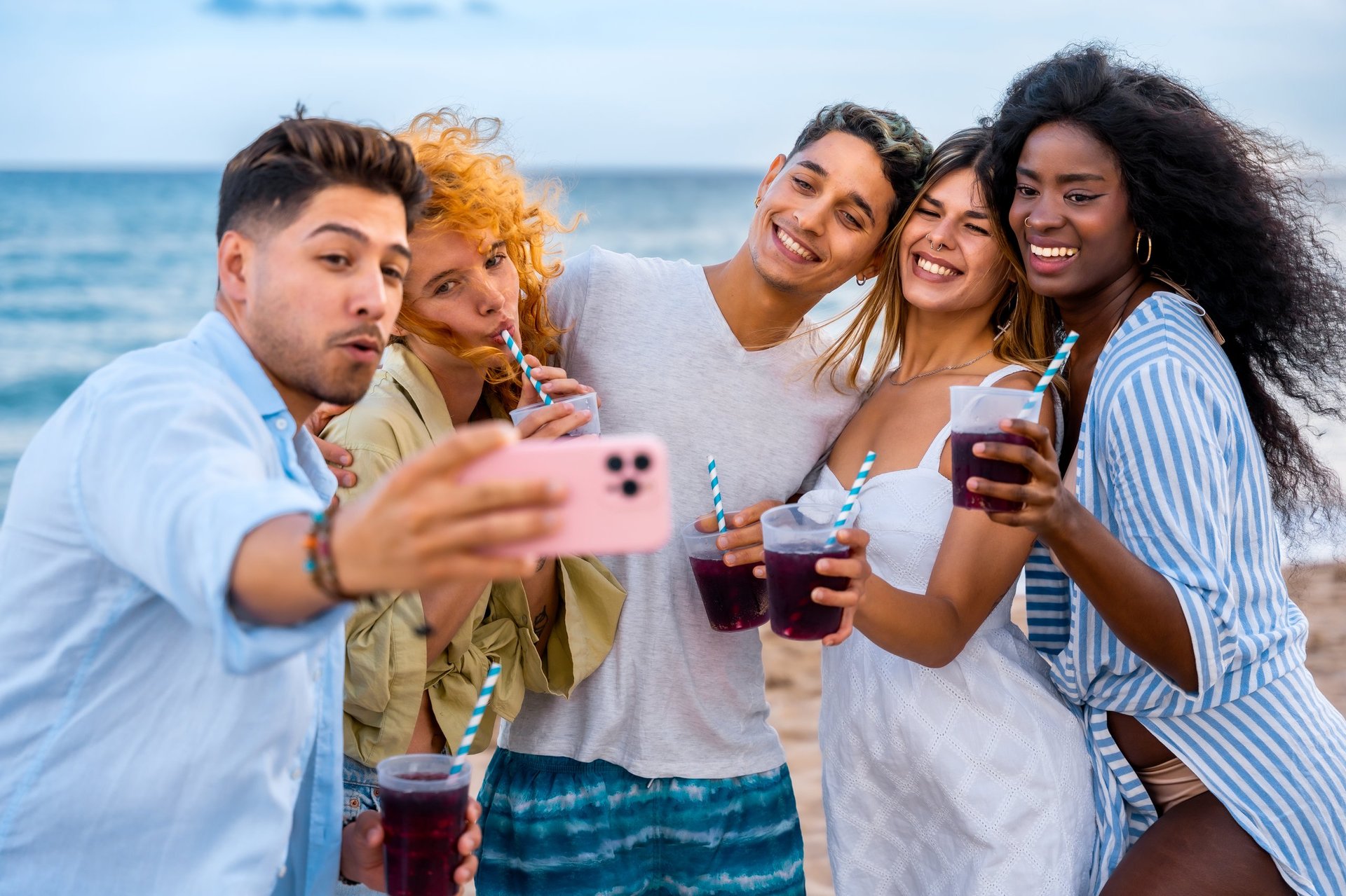 Five cheerful young friends taking a selfie on the beach, enjoying refreshing drinks and the summer vibes