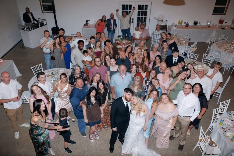 Large group of wedding guests gathered indoors at a reception, with bride and groom in center, dancing and celebrating