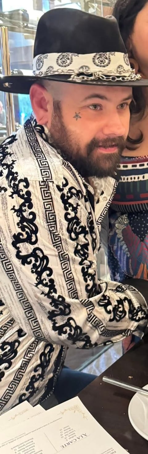 Man with beard and star tattoo wearing ornate black and white patterned jacket and decorative cowboy hat