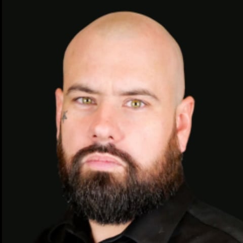 Bald man with full dark beard wearing black shirt against dark background